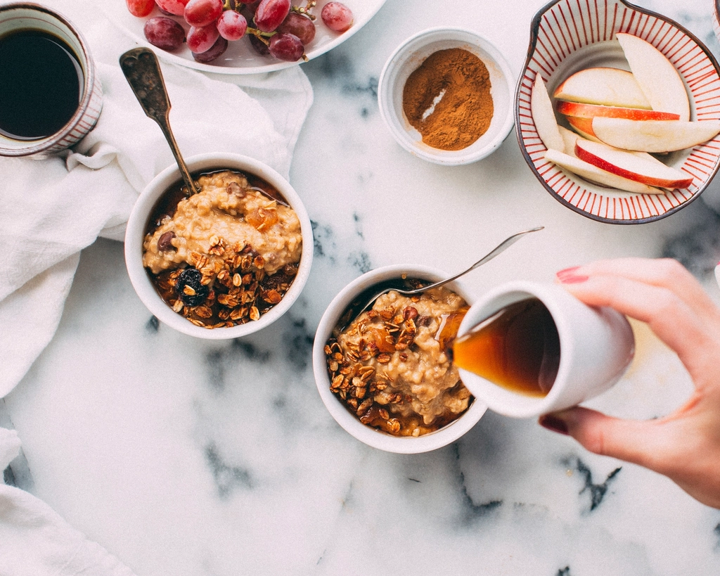A bright overhead flat-lay of two bowls of healthy oatmeal porridge topped with fruit, with fresh berries, sliced apple and a drizzle of honey on a clean marble surface, evoking gut-friendly digestive wellness