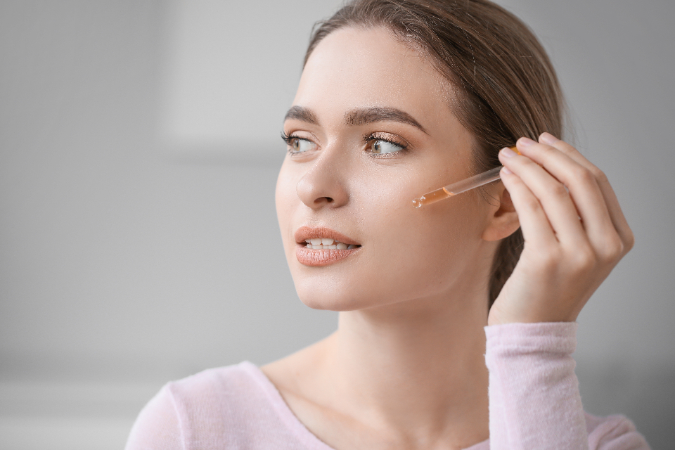A young woman with clear, healthy skin applying a skincare serum to her cheek with a dropper, soft neutral background — evoking a calming skincare routine and fresh, clear complexion.