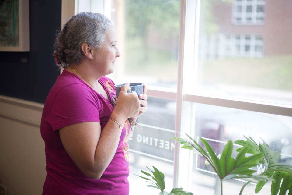 A woman holding a warm mug, standing by a bright window and looking out thoughtfully — a calm, reflective lifestyle moment