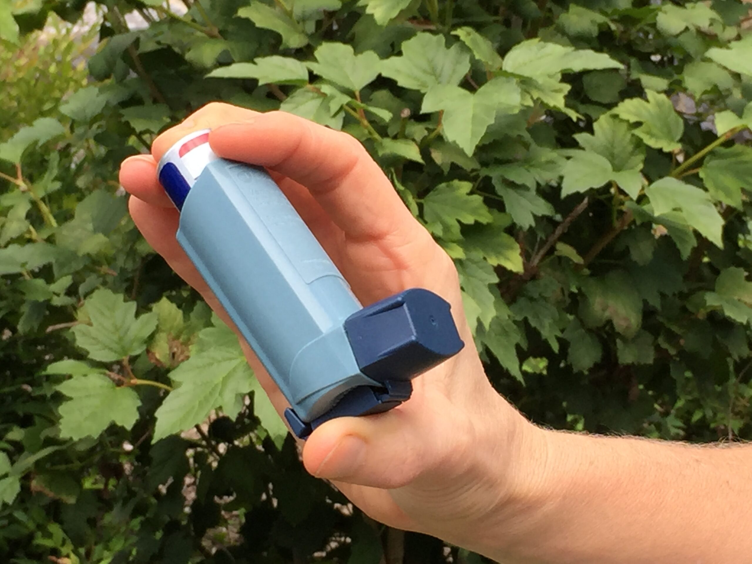 A hand holding a blue asthma reliever inhaler outdoors against lush green foliage, evoking easy breathing and a clean, healthy lifestyle