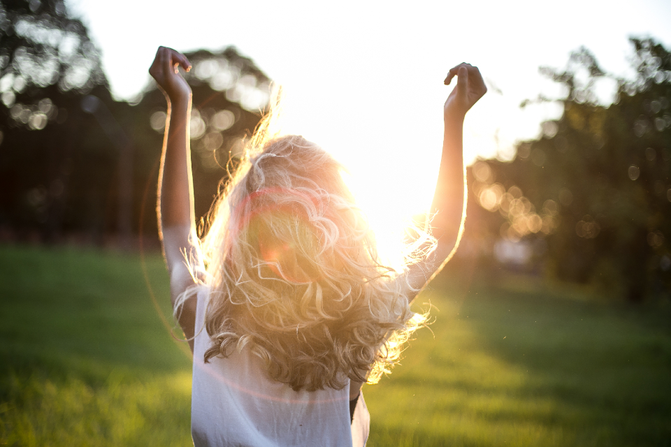 A woman standing in a sunlit green field at golden hour with her arms raised, hair catching the light — a fresh, calm, confident wellness moment.