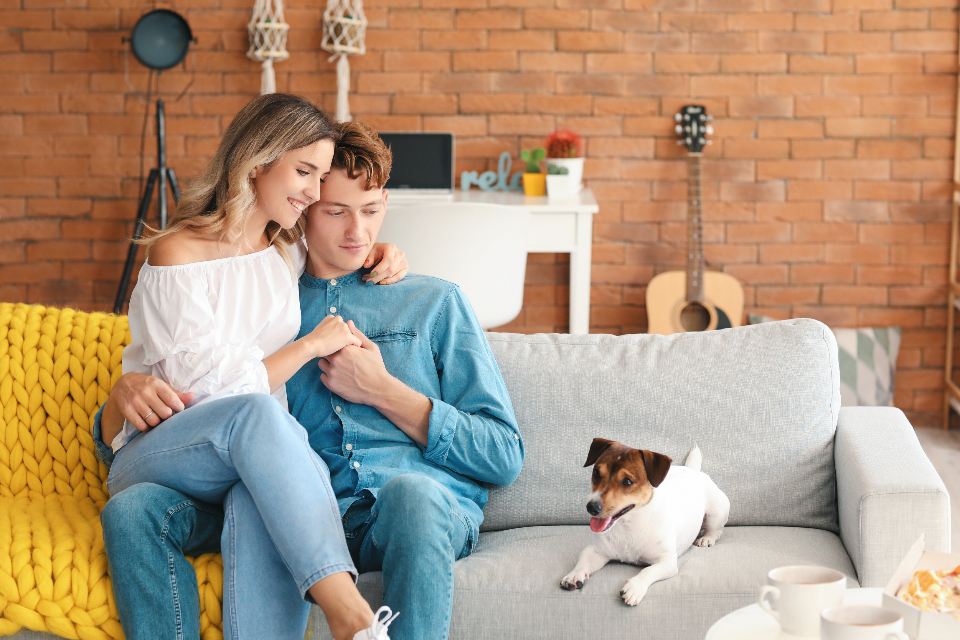 A happy young couple relaxing together at home on a sofa, fully clothed and smiling, conveying reassurance and discreet sexual-health care