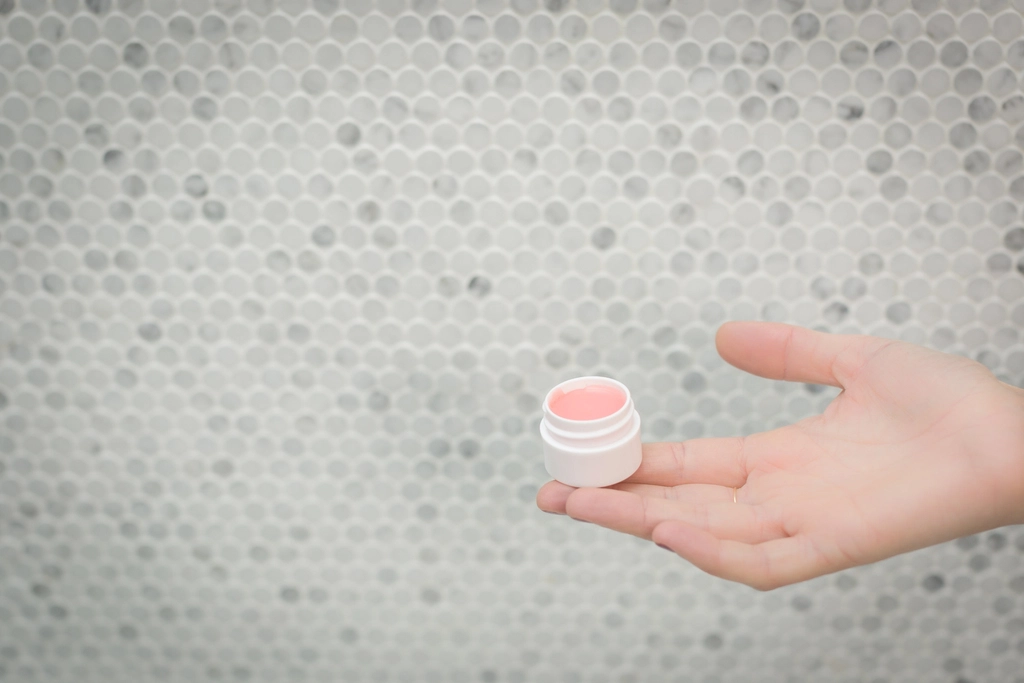 A small open pot of pink lip balm resting in an open palm against a soft, light tiled background, evoking gentle lip care