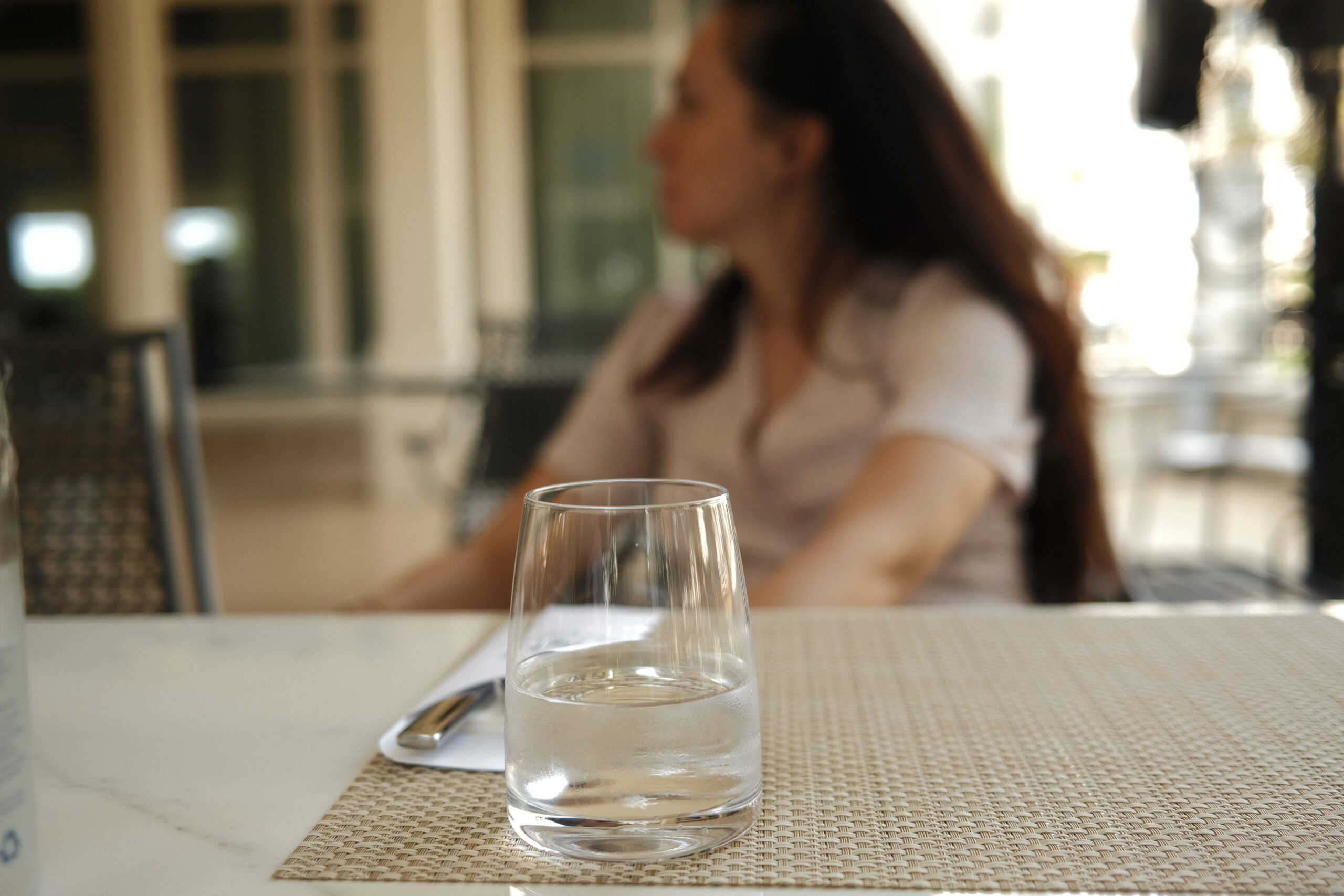 A woman sits relaxed at a sunlit cafe terrace table with a clear glass of water in front of her, evoking calm hydration and everyday wellness.