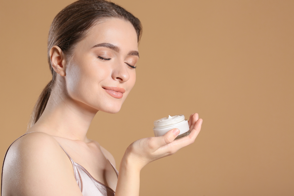 A calm woman with smooth, healthy skin and eyes closed, holding an open jar of moisturising cream against a soft warm background