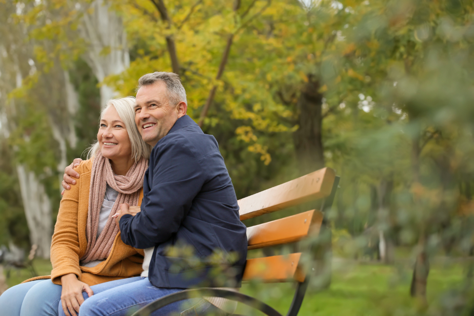 A happy, relaxed mature couple sitting close together on a park bench in autumn, both smiling and looking into the distance