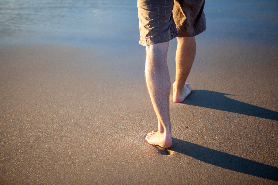 Person walking barefoot with healthy bare feet and legs on warm sand at the shoreline at sunset, evoking clean skin and active foot care