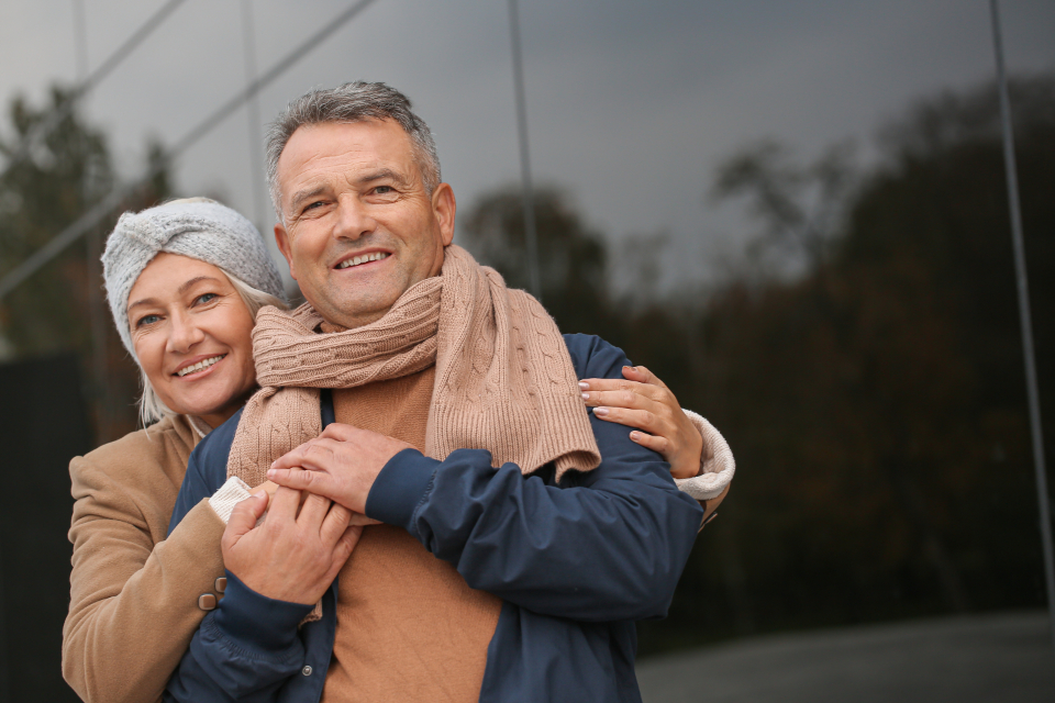A smiling, supportive mature couple embracing warmly outdoors in autumn coats, conveying reassurance, partnership and discreet care.