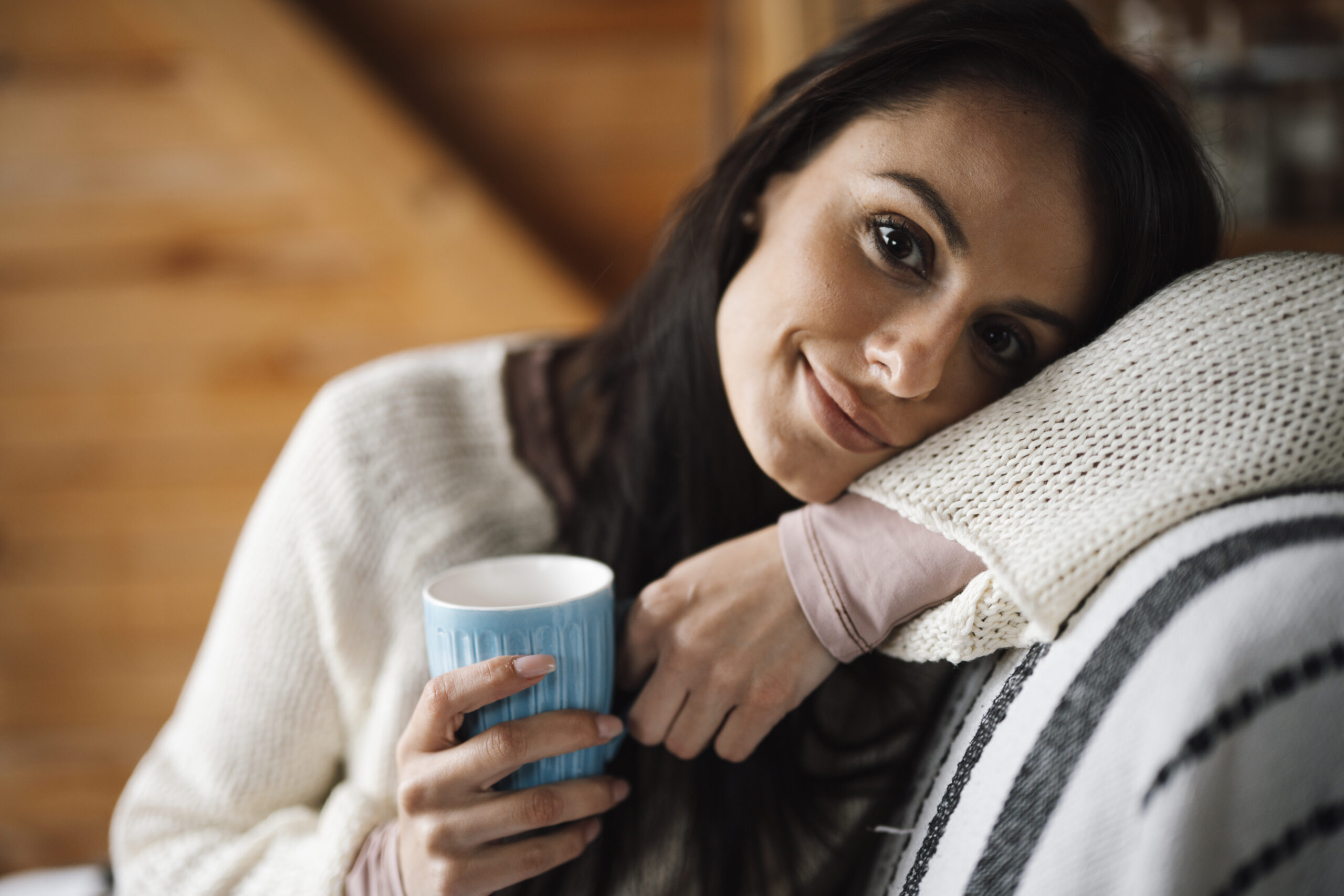 A woman rests comfortably on a sofa, her head leaning on a soft knitted cushion, holding a warm drink and smiling gently in a cosy, calm home setting.
