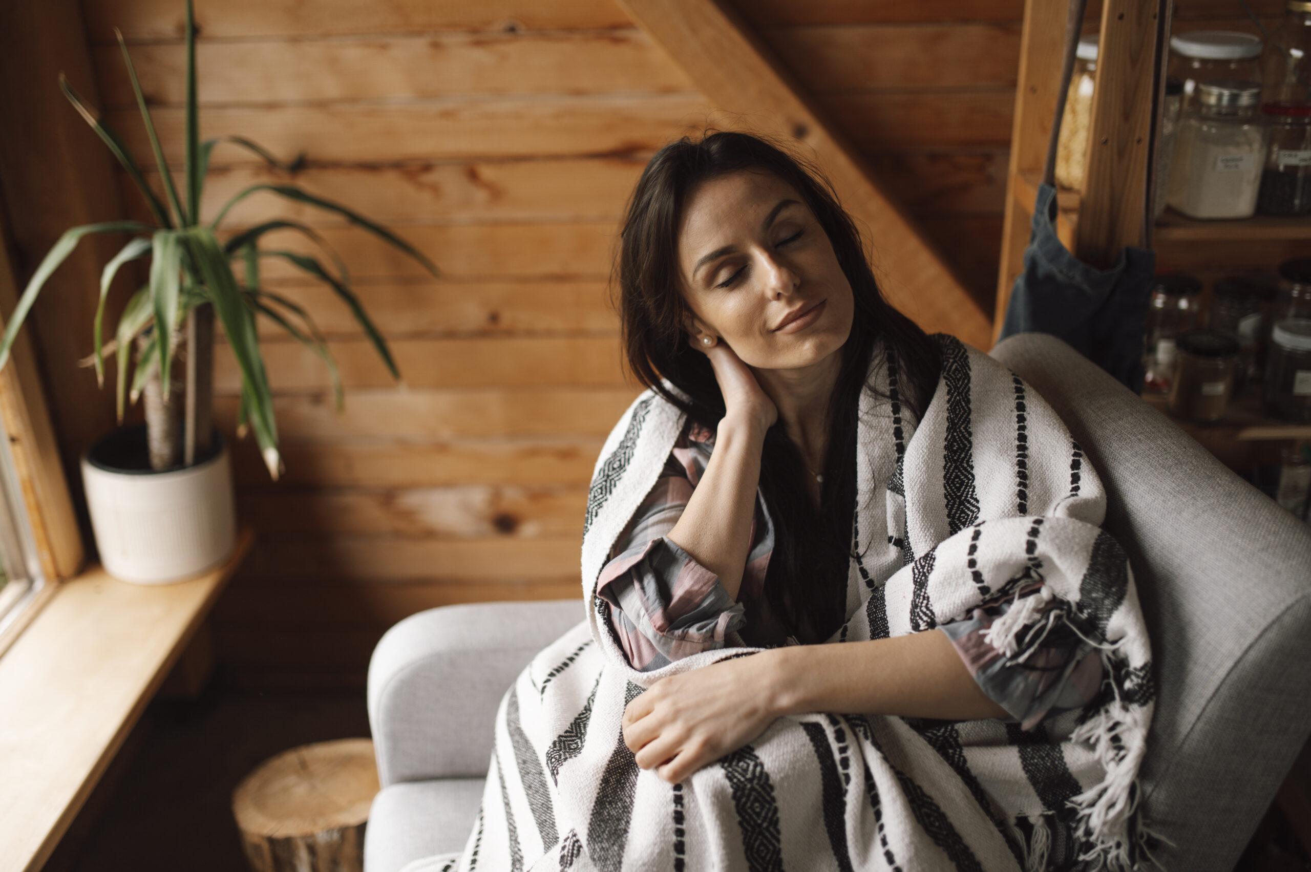 A woman resting with her eyes closed, wrapped in a woven blanket on a sofa in a warm, cosy room — recovering comfortably at home.