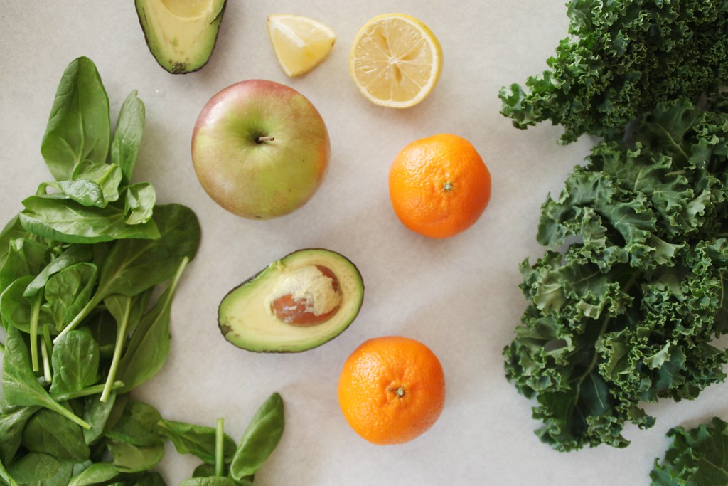 A bright overhead flat-lay of fresh gut-friendly ingredients on a pale surface — spinach, kale, avocado, apple, orange and lemon