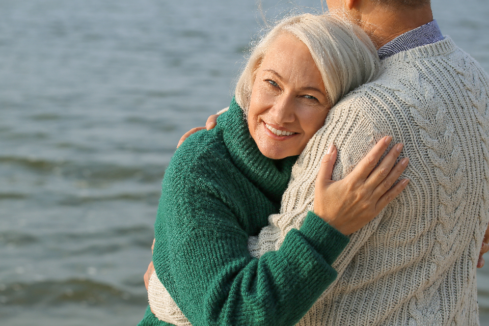 A confident, healthy woman in her fifties with grey hair smiling warmly while embracing her partner outdoors by the water, evoking wellness and vitality in later life.