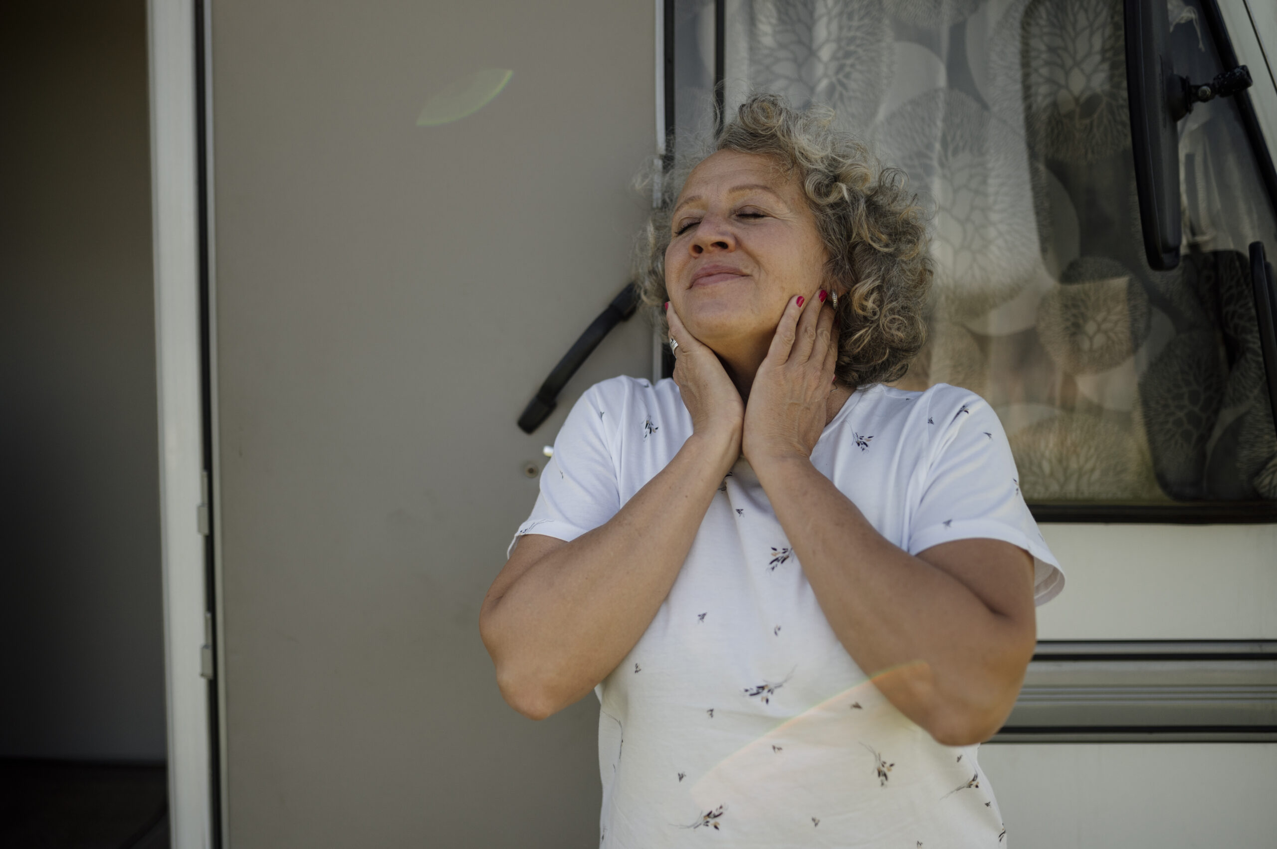 A woman with her eyes closed, hands resting gently against her neck and jaw, expression calm and relieved in soft natural daylight