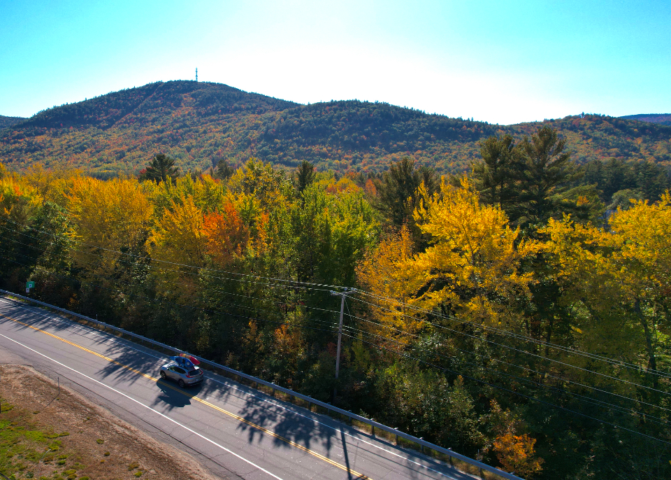 A car driving along a scenic open country road winding through golden autumn forest with rolling mountains under a clear blue sky