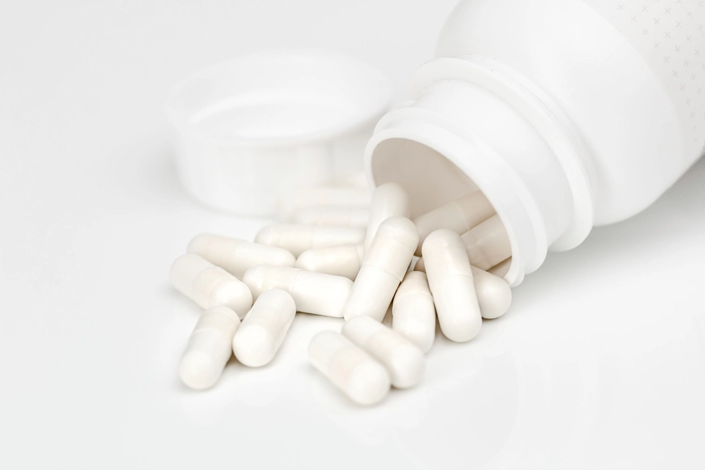 White medication capsules spilling from an open white pill bottle on a clean, bright background, representing a prescribed course of antibiotic treatment.