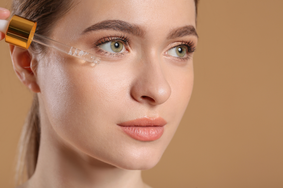 Woman with clear, healthy skin applying a moisturising skincare serum with a dropper, against a soft warm beige background