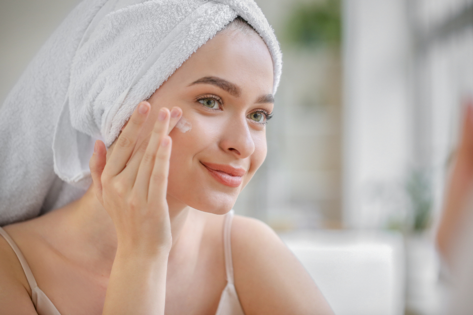Smiling young woman with a fresh towel turban gently applying skincare to her glowing, clear cheek in a bright, airy bathroom