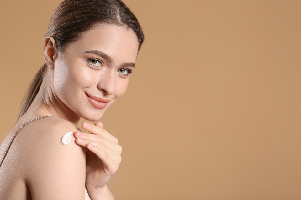 Young woman with clear, healthy glowing skin applying moisturising cream to her shoulder against a warm neutral background
