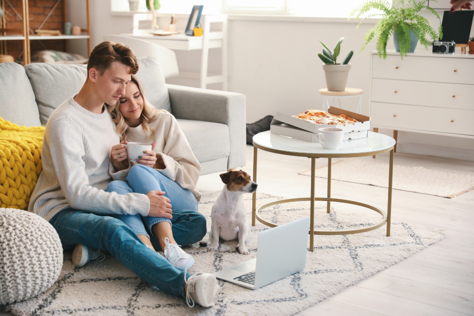 A relaxed young couple in casual knitwear and jeans sitting together on the floor of a bright, tidy living room with a coffee mug and their dog, looking calm and reassured.