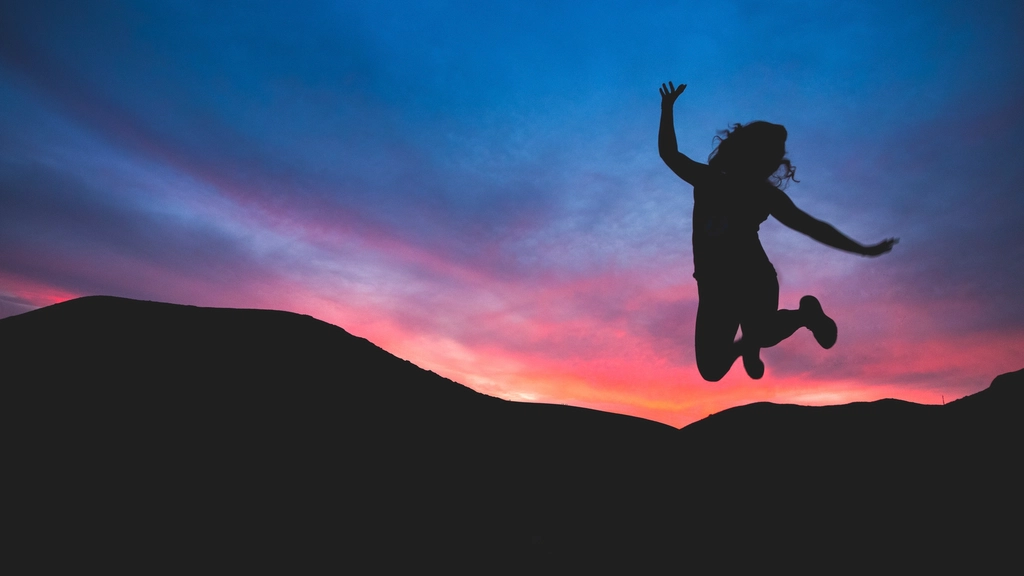 Silhouette of a woman leaping joyfully with arms raised against a vivid pink and blue sunset sky over rolling hills, evoking freedom and renewed vitality after quitting smoking