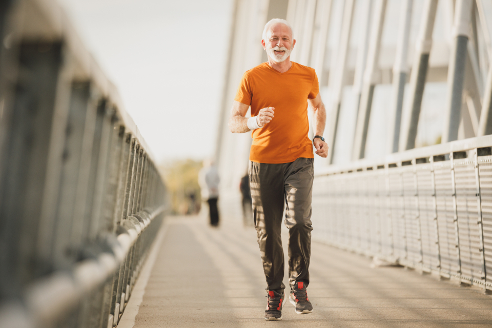 Fit, smiling mature man with grey hair and beard jogging across a bridge in athletic wear, conveying energy and vitality